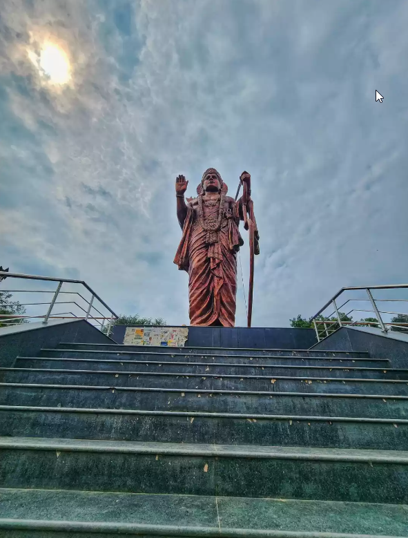 Tapovan Development Debate Nashik – statue of a revered saint standing atop stone steps under a dramatic cloudy sky.